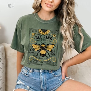 K&ouml;nnte beinhalten: Gr&uuml;nes T-Shirt mit kurzen &Auml;rmeln und einem Vintage-Motiv einer Biene und einer Sonnenblume. Der Schriftzug lautet "Bee Kind, Bee Positive, Bee Humble, Bee Honest, Bee Grateful, Believe in Yourself."