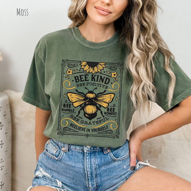 K&ouml;nnte beinhalten: Gr&uuml;nes T-Shirt mit kurzen &Auml;rmeln und einem Vintage-Motiv einer Biene und einer Sonnenblume. Der Schriftzug lautet "Bee Kind, Bee Positive, Bee Humble, Bee Honest, Bee Grateful, Believe in Yourself."