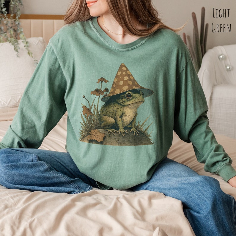 Pode incluir: Uma camisa de manga comprida verde claro com um gr&aacute;fico de um sapo usando um chap&eacute;u de cogumelo marrom e branco. O sapo est&aacute; sentado em uma rocha com plantas verdes ao redor.