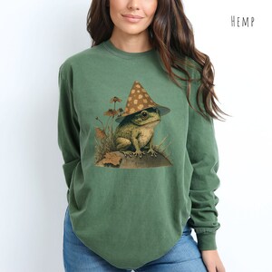Pode incluir: Uma camisa verde de manga comprida com um gr&aacute;fico de um sapo usando um chap&eacute;u marrom e branco com manchas. O sapo est&aacute; sentado em uma rocha com plantas verdes ao redor. A camisa tem a palavra "HEMP" impressa no canto superior direito.