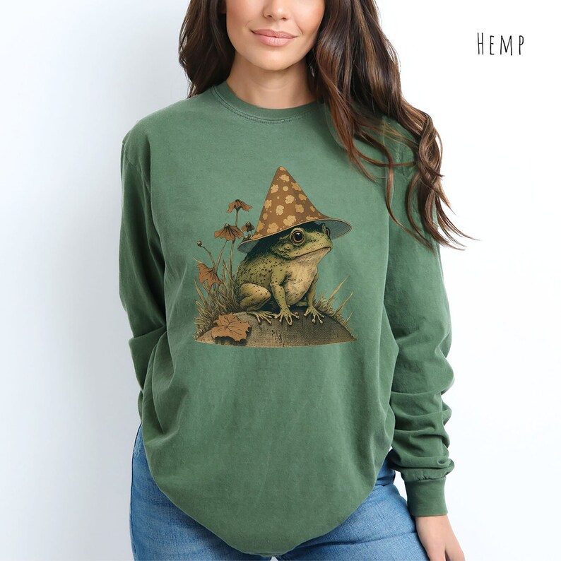 Pode incluir: Uma camisa verde de manga comprida com um gr&aacute;fico de um sapo usando um chap&eacute;u marrom e branco com manchas. O sapo est&aacute; sentado em uma rocha com plantas verdes ao redor. A camisa tem a palavra "HEMP" impressa no canto superior direito.