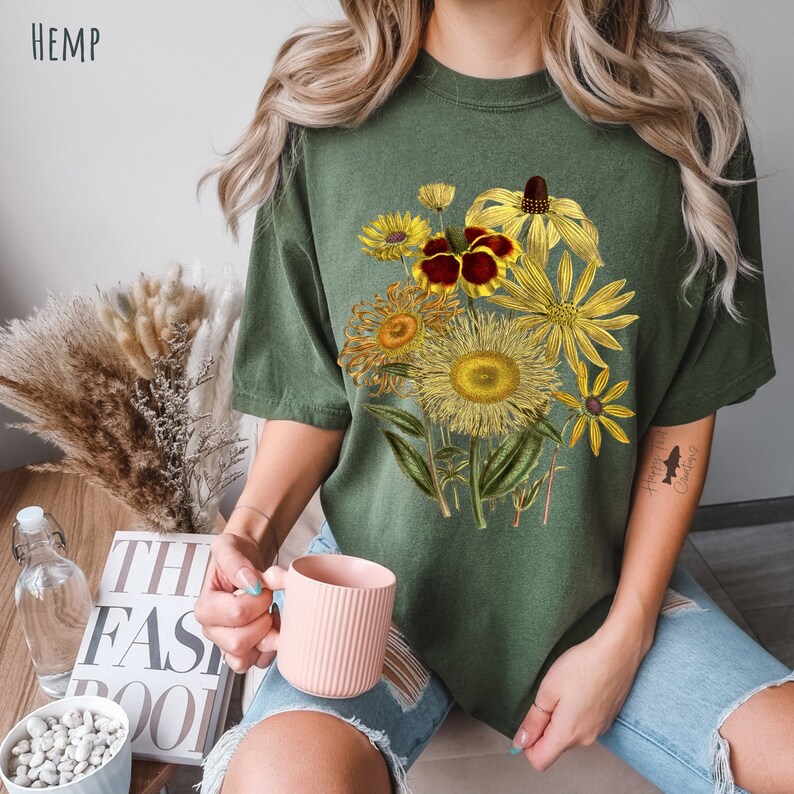 Op de afbeelding: Een vrouw draagt een groen T-shirt met een bloemenprint van gele, rode en bruine bloemen. Het shirt heeft het woord "HEMP" linksboven gedrukt.