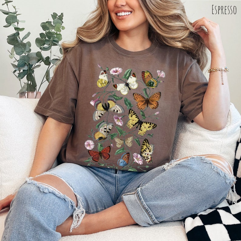 May include: A brown t-shirt featuring a vintage illustration of butterflies and flowers. The butterflies are in various colours, including yellow, orange, red, and blue.