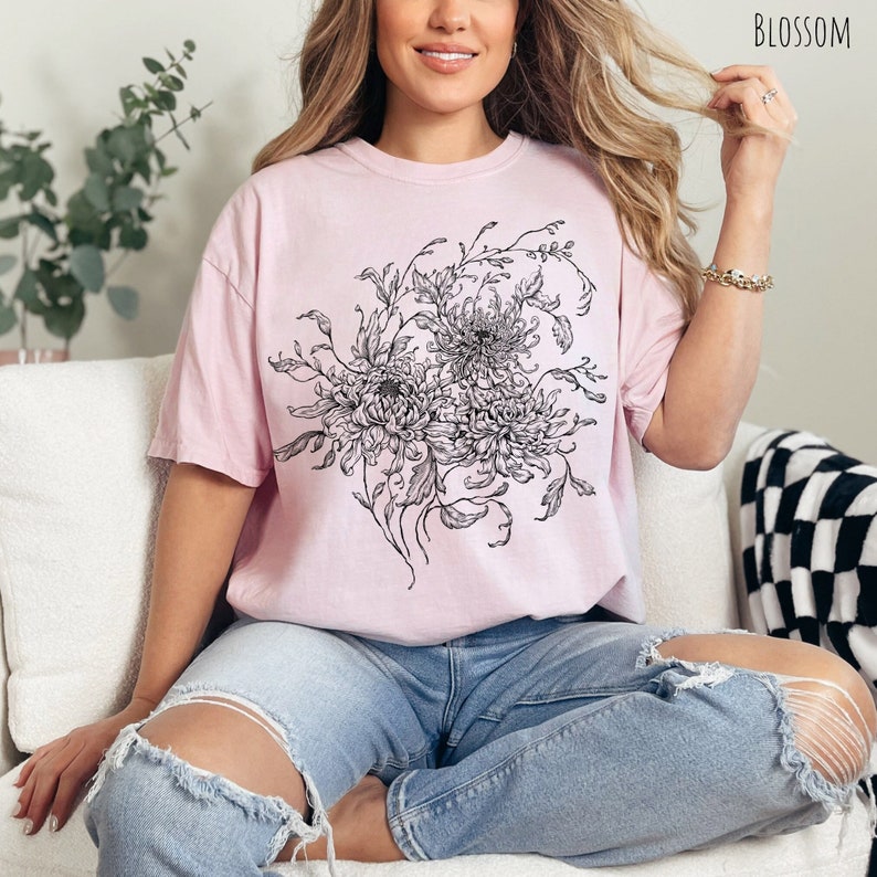 May include: An oversized pink t-shirt featuring a black and white floral print of chrysanthemums. The shirt has a relaxed fit and a crew neckline.