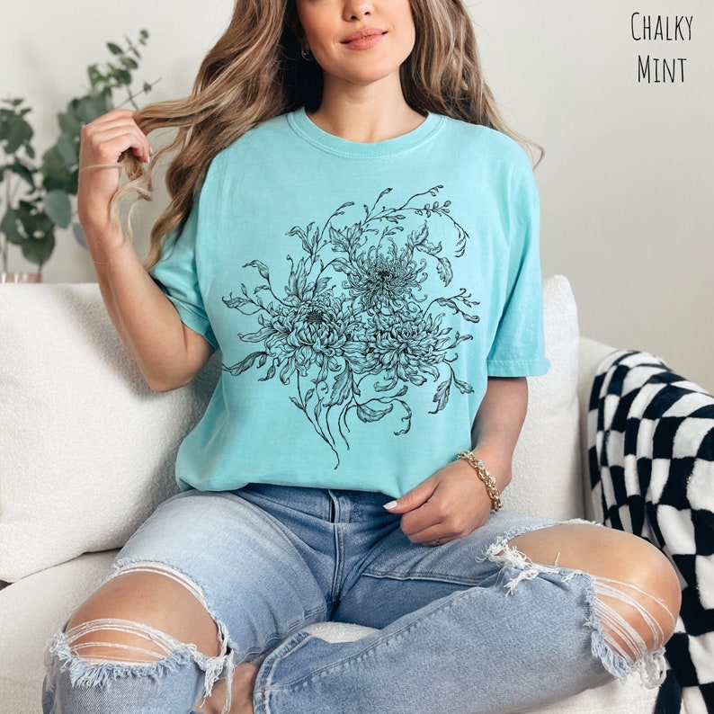 May include: A light blue t-shirt featuring a black and white floral design. The design depicts a bouquet of flowers with intricate details. The text "Chalky Mint" is visible in the top right corner of the image.