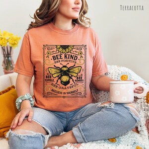 K&ouml;nnte beinhalten: Ein terracottafarbenes T-Shirt mit einer Grafik im Vintage-Stil einer Biene und einer Sonnenblume. Die Grafik lautet "Bee Kind. Bee Positive. Bee Humble. Bee Honest. Bee Grateful. Believe in Yourself."