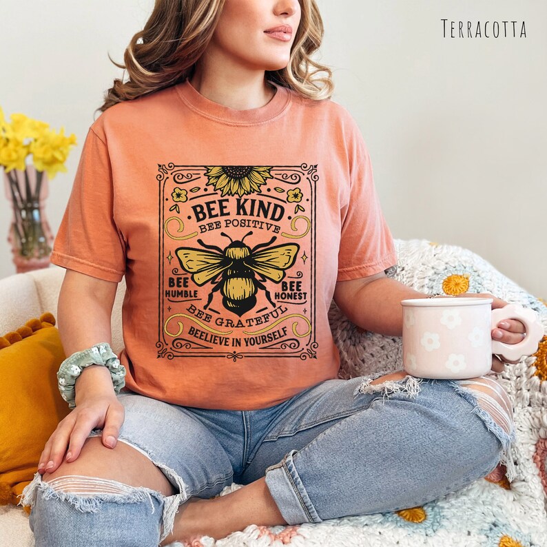 K&ouml;nnte beinhalten: Ein terracottafarbenes T-Shirt mit einer Grafik im Vintage-Stil einer Biene und einer Sonnenblume. Die Grafik lautet "Bee Kind. Bee Positive. Bee Humble. Bee Honest. Bee Grateful. Believe in Yourself."