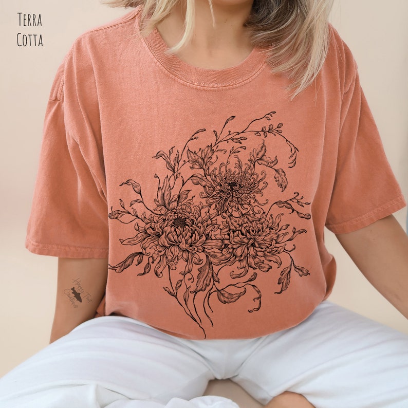 May include: A terracotta coloured t-shirt with a black and white floral design featuring chrysanthemums. The design is centred on the shirt and covers most of the front.