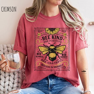 K&ouml;nnte beinhalten: Ein rotes T-Shirt mit einer Grafik im Vintage-Stil, die eine Biene und den Text "Bee Kind. Bee Positive. Bee Humble. Bee Honest. Bee Grateful. Beelieve in Yourself." zeigt.