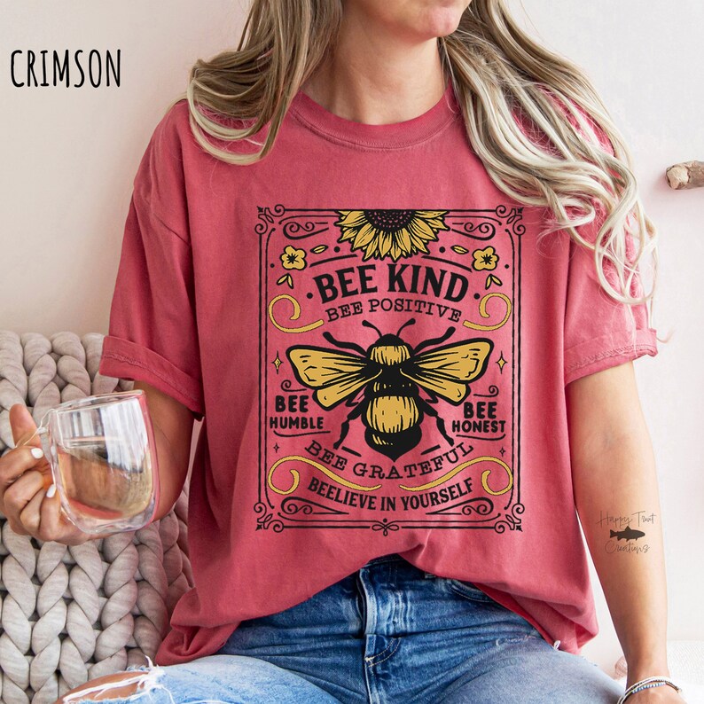 K&ouml;nnte beinhalten: Ein rotes T-Shirt mit einer Grafik im Vintage-Stil, die eine Biene und den Text "Bee Kind. Bee Positive. Bee Humble. Bee Honest. Bee Grateful. Beelieve in Yourself." zeigt.