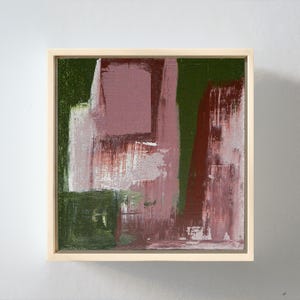 May include: A square canvas painting featuring abstract brushstrokes in shades of deep forest green, muted mauve, and dark burgundy. The artwork is encased in a light-colored wooden frame, displaying a textured application of paint across the surface.