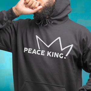 May include: Dark gray hoodie with a white crown graphic and the text "PEACE KING." The hoodie has a drawstring hood and a front pocket. The design is simple and bold, suitable for casual wear.