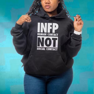 May include: Black hoodie with the text "INFP HUMAN CONTACT NOT SOCIAL CONTACT" in white. The hoodie has a front pocket and a drawstring hood. The person in the image is wearing blue jeans.