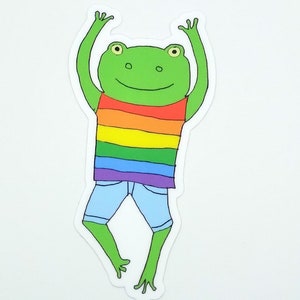 May include: A green frog wearing a rainbow striped shirt and blue denim shorts with its arms raised in the air.