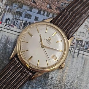 May include: A vintage gold-toned Omega Automatic wristwatch with a light-coloured face, gold markers, and a date window at the 3 o'clock position. The watch has a brown leather strap. The watch face displays the Omega logo.
