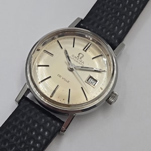 May include: A vintage Omega De Ville automatic wristwatch with a silver dial and black leather strap. The watch face displays the date and the Omega logo. The watch has a silver case and a textured black leather band.