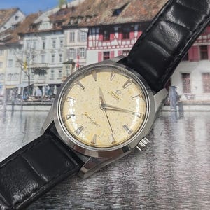 May include: A vintage Omega Seamaster automatic wristwatch with a cream-colored speckled dial and silver hands. The watch has a stainless steel case and a black leather strap. The background is a blurred image of a European town.