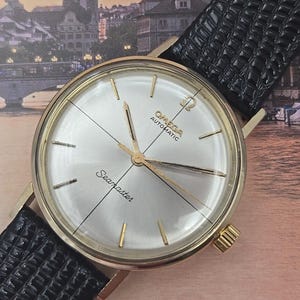 May include: A gold-toned Omega Seamaster automatic wristwatch with a silver dial and gold hands and markers. The watch has a black leather band and a gold crown. The watch face displays the Omega logo and the word "Seamaster".