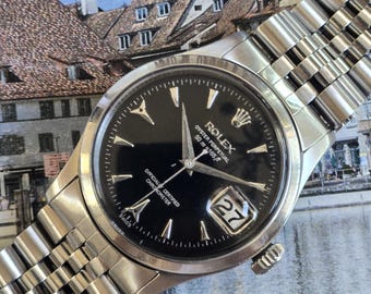 May include: A stainless steel Rolex Oyster Perpetual wristwatch with a black dial, silver-tone hands, and a date window displaying the number 27. The watch features a multi-link metal bracelet and text reading Rolex Oyster Perpetual 50m=165ft.