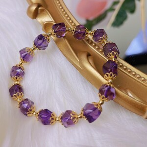 Pode incluir: Uma pulseira dourada com contas de ametista roxa profunda facetadas. As contas são separadas por detalhes ornamentados em filigrana dourada. A pulseira é exibida em uma superfície dourada, com um fundo branco macio.