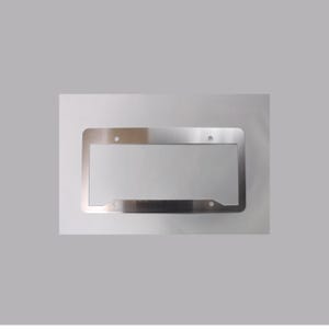 May include: A rectangular, metallic silver license plate frame with a smooth, reflective finish. The frame features four circular mounting holes, two positioned along the top edge and two along the bottom edge, designed for standard vehicle attachment.