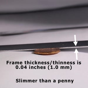 May include: A thin, flat, dark-colored frame edge positioned above a copper penny. White arrows indicate the frame thickness, which is labeled as 0.04 inches (1.0 mm). The text below states: Frame thickness/thinness is 0.04 inches (1.0 mm). Slimmer than a penny.