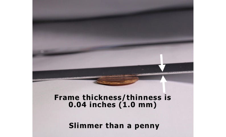 May include: A thin, flat, dark-colored frame edge positioned above a copper penny. White arrows indicate the frame thickness, which is labeled as 0.04 inches (1.0 mm). The text below states: Frame thickness/thinness is 0.04 inches (1.0 mm). Slimmer than a penny.