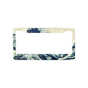May include: A rectangular license plate frame featuring a printed design of a large, stylized blue ocean wave with white foam crests against a cream-colored background. The frame includes four circular mounting holes at the corners for attachment.