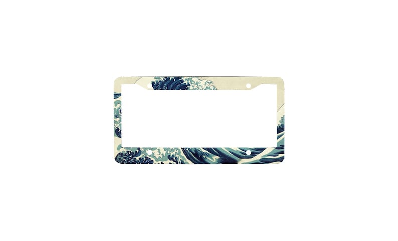 May include: A rectangular license plate frame featuring a printed design of a large, stylized blue ocean wave with white foam crests against a cream-colored background. The frame includes four circular mounting holes at the corners for attachment.