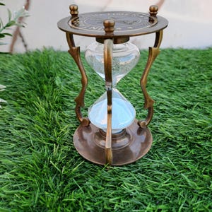 Könnte beinhalten: Eine Sanduhr aus Messing mit einem verzierten Sockel und einer runden Oberseite mit einer Inschrift. Die Sanduhr ist mit weißem Sand gefüllt und steht auf einer grünen Oberfläche.