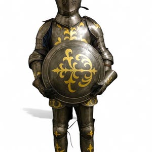 Peut inclure: Une armure médiévale complète dans les tons de gris et d'or. L'armure comprend un casque, une cuirasse, des protège-bras et de jambes, et un bouclier. Le bouclier est rond et présente un motif floral doré. L'armure est conçue pour protéger le porteur.