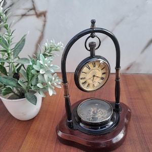 May include: A vintage desk clock and compass. The clock face is gold with black Roman numerals and the text "VICTORIA LONDON 1876". The clock and compass are set in a black metal frame with a wooden base.