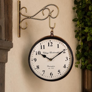 May include: A round, double-sided wall clock with a dark metal frame and a cream-colored face. The dial features black Arabic numerals, black hands, and the text Village Clockworks, Birmingham, est. 1792. It hangs from a decorative brass-toned scroll bracket.