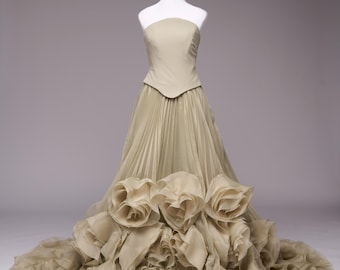 May include: A strapless, pale green gown on a white mannequin. The bodice is fitted, and the skirt features pleated fabric and large, ruffled floral accents at the hem. The dress is displayed against a neutral background.