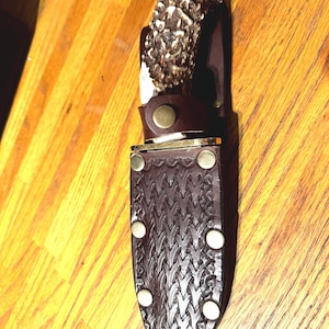 May include: A handcrafted hunting knife with a deer antler handle and a brown leather sheath. The sheath features a decorative embossed pattern and is secured with brass rivets and a leather strap.