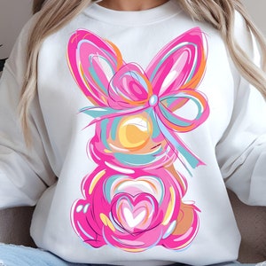 May include: White sweatshirt with a pink and blue abstract bunny design. The bunny has a large pink bow and a heart in the center of its chest.