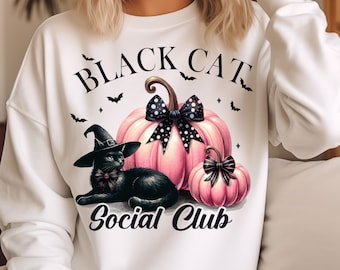 May include: White sweatshirt with a black cat wearing a witch's hat and a pink pumpkin with black polka dots and a black bow. The text "Black Cat Social Club" is printed on the sweatshirt.