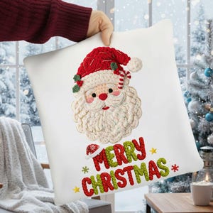 May include: White square Christmas cushion featuring a detailed Santa Claus face with a red hat and beard. The words "MERRY CHRISTMAS" are embroidered in red and green. The cushion is held up in front of a snowy window.