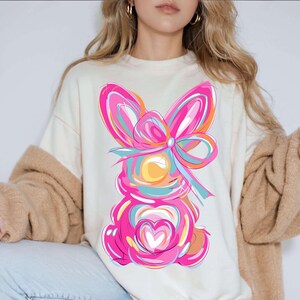 Peut inclure: Un sweat-shirt blanc avec un motif color&eacute; d'un lapin avec un n&oelig;ud rose. Le lapin tient un c&oelig;ur dans ses pattes.