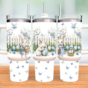 Op de afbeelding: Drie witte reisbekers met roestvrijstalen rietjes en handvatten. Elke beker heeft een aquarelontwerp van paashazen, eieren en een wit hek met blauwe bloemaccenten en vlinders. De bekers staan op een houten oppervlak.