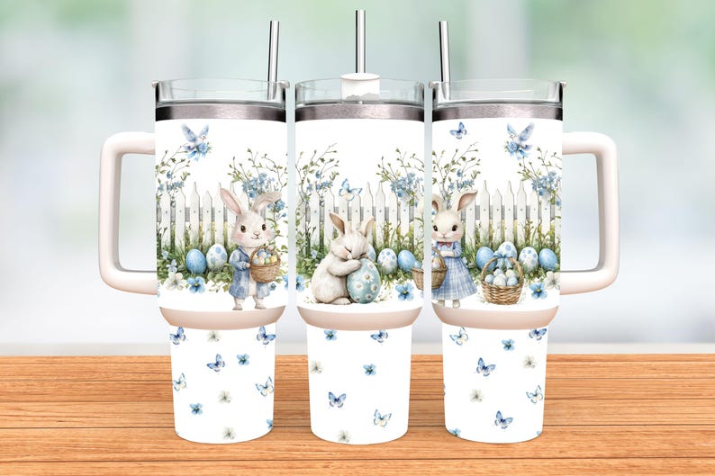 Op de afbeelding: Drie witte reisbekers met roestvrijstalen rietjes en handvatten. Elke beker heeft een aquarelontwerp van paashazen, eieren en een wit hek met blauwe bloemaccenten en vlinders. De bekers staan op een houten oppervlak.
