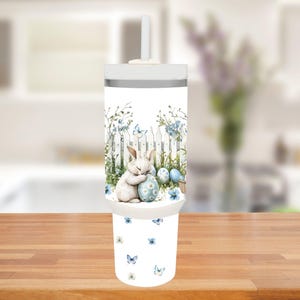 Op de afbeelding: Een witte beker met deksel en rietje, met een Paasthema. Het ontwerp toont een slapend konijn dat een ei omarmt, een witte hek, blauwe bloemen en vlinders. De onderkant van de beker heeft een patroon van blauwe vlinders en bloemen.