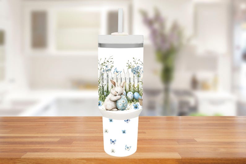 Op de afbeelding: Een witte beker met deksel en rietje, met een Paasthema. Het ontwerp toont een slapend konijn dat een ei omarmt, een witte hek, blauwe bloemen en vlinders. De onderkant van de beker heeft een patroon van blauwe vlinders en bloemen.