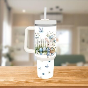 Op de afbeelding: Een witte beker met een handvat en een rietje, met een paasthema. Het ontwerp bevat een konijn, eieren, bloemen en vlinders in blauw en wit. De beker staat op een houten oppervlak.