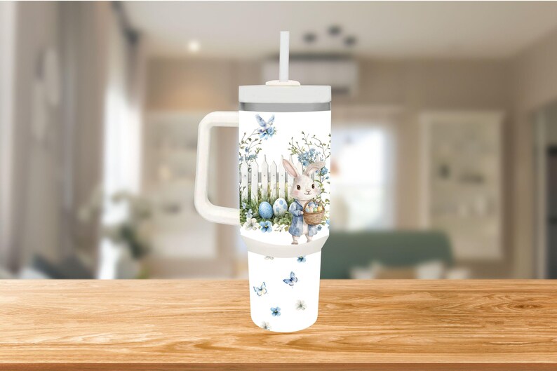 Op de afbeelding: Een witte beker met een handvat en een rietje, met een paasthema. Het ontwerp bevat een konijn, eieren, bloemen en vlinders in blauw en wit. De beker staat op een houten oppervlak.