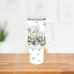 Op de afbeelding: Witte beker met een handvat en een rietje, met een Paasontwerp. Het ontwerp toont een konijn in een blauwe jurk, met manden met eieren, tegen een witte hek en blauwe bloemaccenten. Vlinders zijn verspreid.