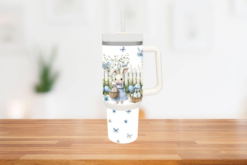Op de afbeelding: Witte beker met een handvat en een rietje, met een Paasontwerp. Het ontwerp toont een konijn in een blauwe jurk, met manden met eieren, tegen een witte hek en blauwe bloemaccenten. Vlinders zijn verspreid.
