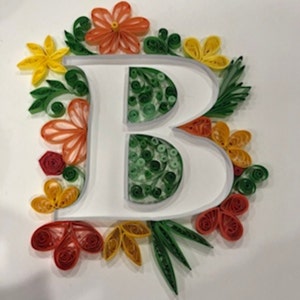 Può includere: Un'opera d'arte in quilling di carta bianca con la lettera "B" circondata da colorati fiori e foglie di carta. I fiori sono nelle tonalità del giallo, arancione e rosso, con foglie verdi. La lettera è riempita di quilling di carta verde.