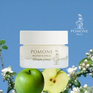 May include: A white jar of Pomone Munificence eye serum with a gold label and a green apple in the foreground. The jar is on a white background with a blue sky and white flowers.