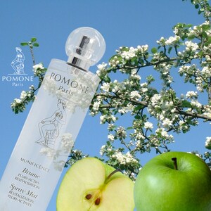 May include: A clear glass bottle of POMONE Paris Brume Spray Mist with a white label and a gold logo. The bottle is sitting on a green apple and a half apple. The background is a blue sky with white flowers.